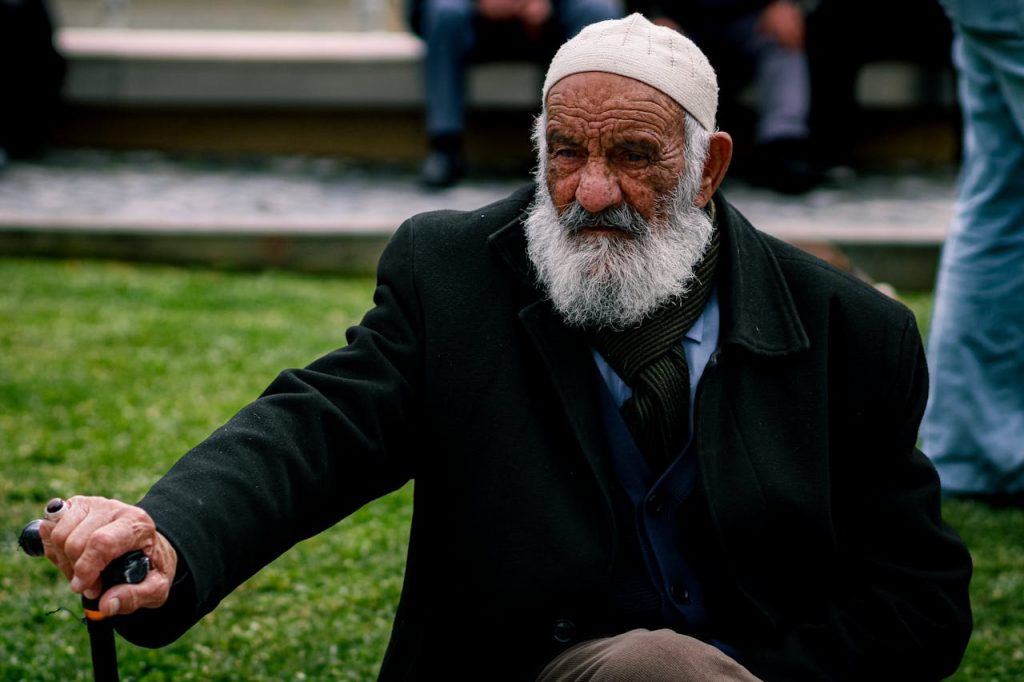 Portrait of an elderly man in a park sitting with a cane, wearing a white cap and a thoughtful expression.
